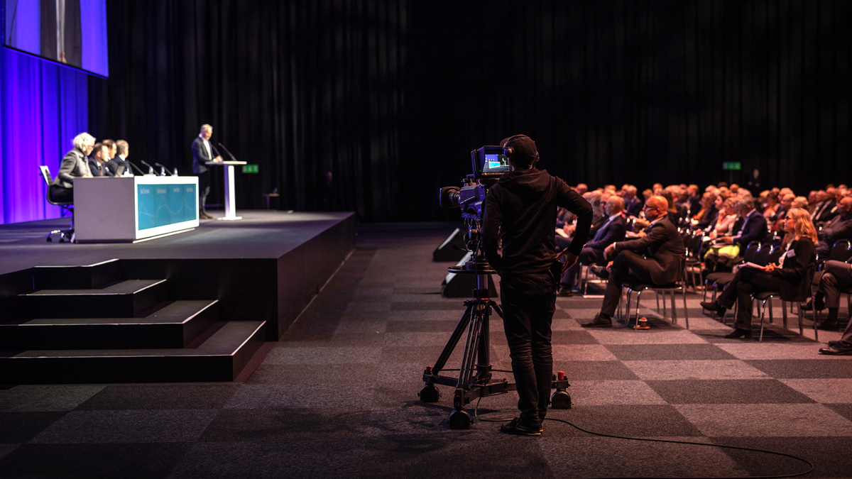 The presenters at a AGM sitting on a stage in front of a large audience below and the back of a person with camera equipment filming the event