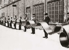 Historical photo of a line of about 20 men holding a long strip steel outside a factory building.