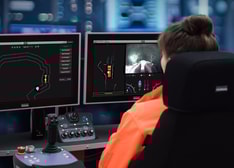 A woman sitting at a desk in a control room looking into two monitors showing different views of a mine