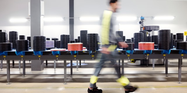 A man in work clothes passing a long bench with trays with inserts on it in an industrial setting