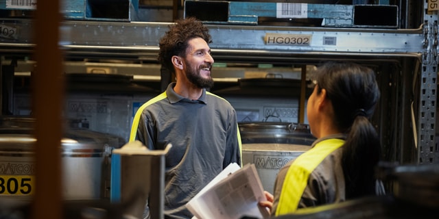 A man and a woman wearing work clothes facing each other in an industrial environment