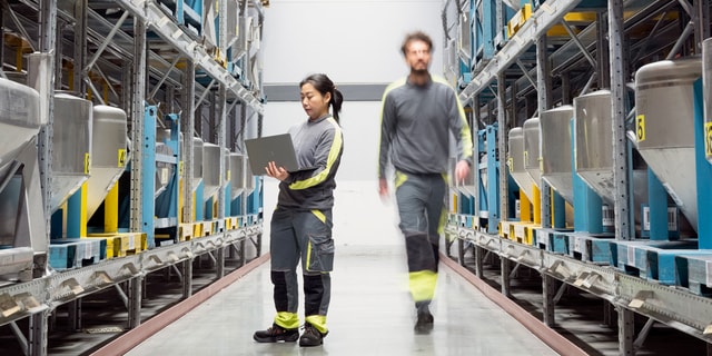 A woman holding a laptop and a man walking between two warehouse shelfs