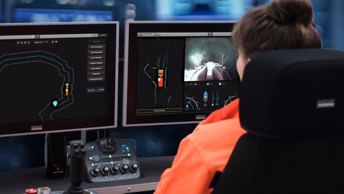 A woman sitting at a desk in a control room looking into two monitors showing different views of a mine