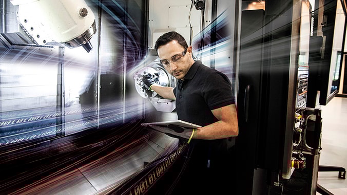 A man standing next to a CNC machine looking on a tabket that he holds in one of his hands