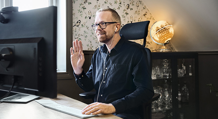 A man with earphones sitting in a home environment at a desk waving towards the computer screen