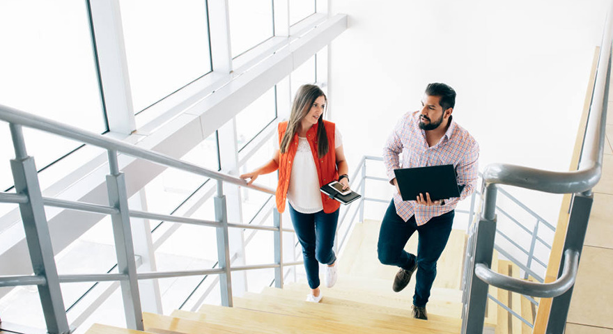 A woman holding a phone and a notebook and a man holding a laptop while they walk beside each other up a stair