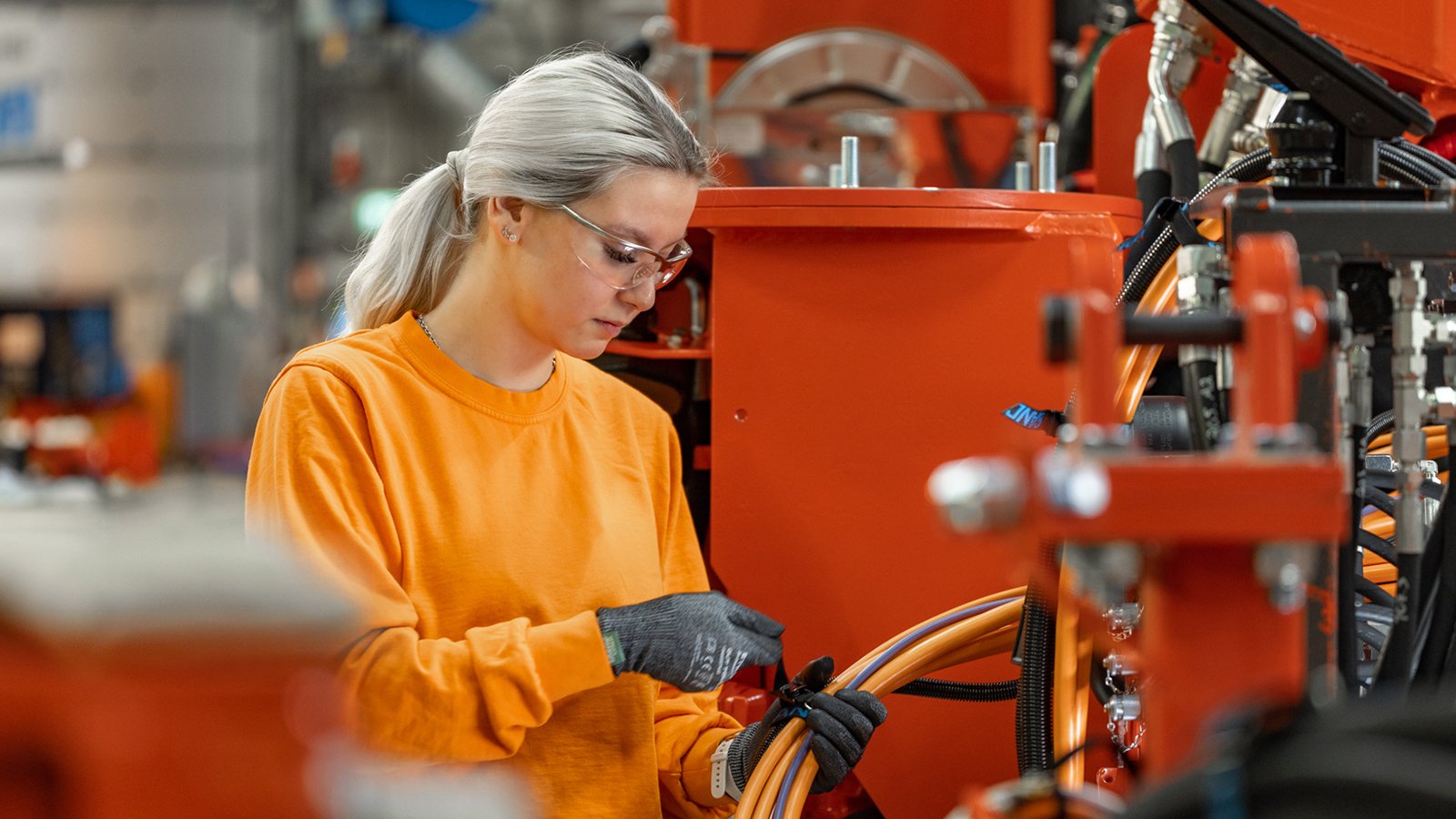 A woman in work clothes putting together machine cables in an industrial environment. 