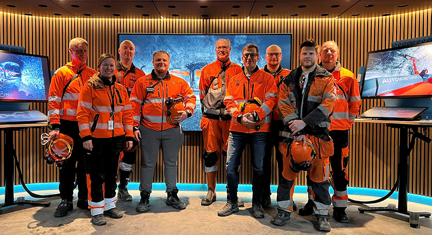 Nine people wearing personal protective equipment for mining operations standing next to each other with three large monitors behind them.