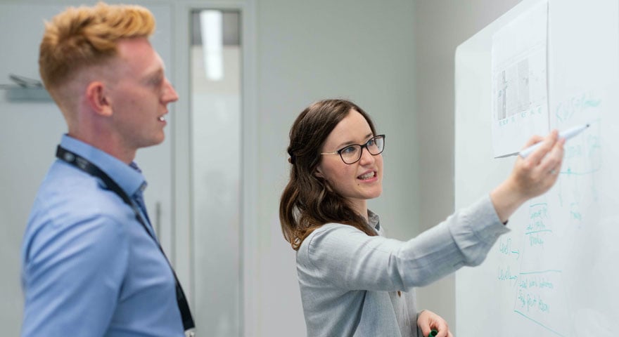 A woman and a man in front of a whiteboard, the woman writing on it