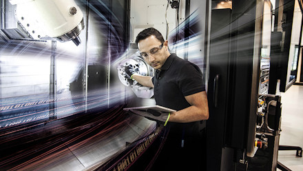 A man standing next to a CNC machine looking on a tabket that he holds in one of his hands