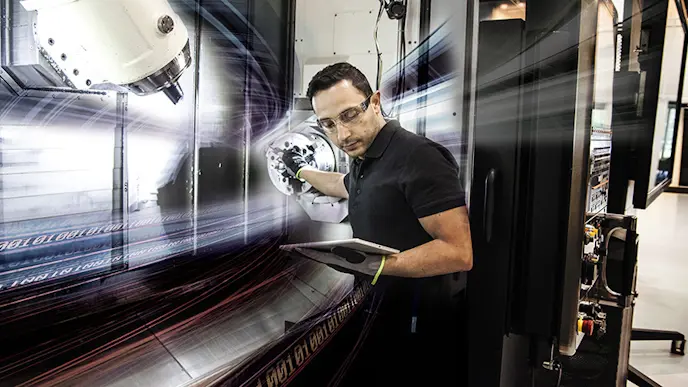 A man standing next to a CNC machine looking on a tabket that he holds in one of his hands
