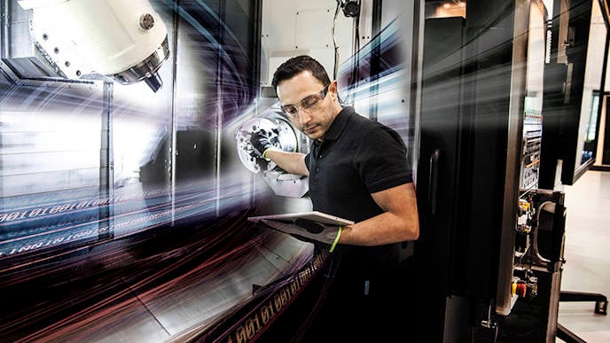 A man standing next to a CNC machine looking on a tabket that he holds in one of his hands