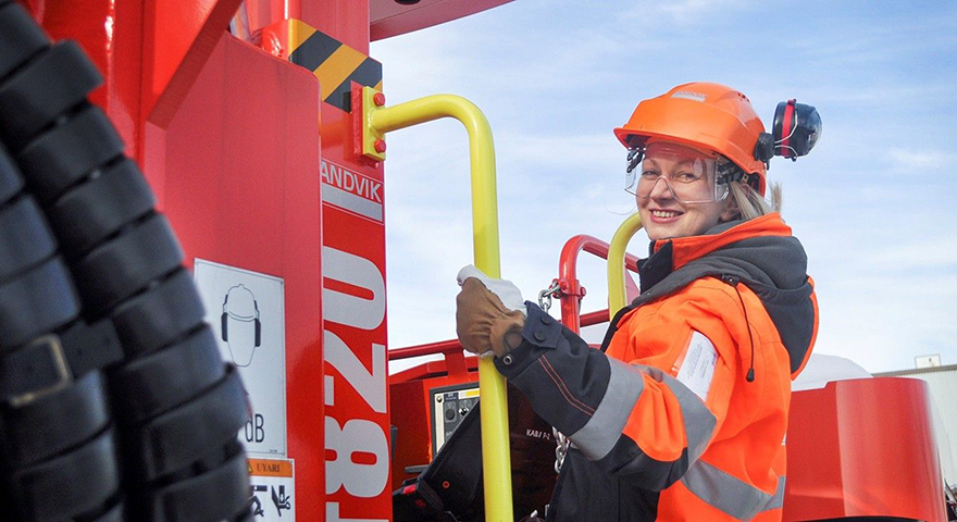 A woman wearing orange work clothes and a helmet steps up into a Sandvik tunneling jumbo.